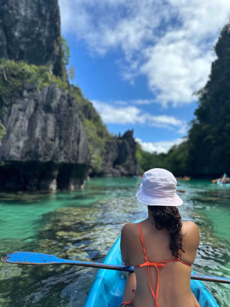 Solo female traveler kayaking through Big Blue Lagoon in El Nido, Philippines while wearing an orange swim suit and white bucket hat