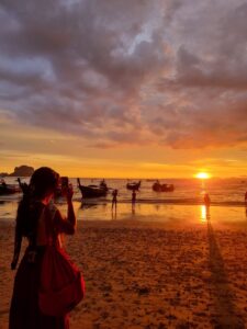 Solo female traveler taking a picture of the longtail boats and sunset on railay beach thailand