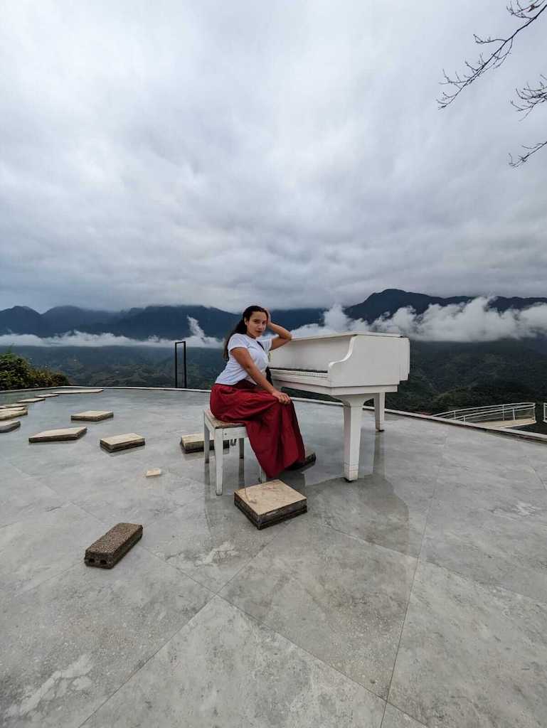 Solo female traveler sitting next to a white piano in Moana Sapa Vietnam while wearing a white skirt and red skirt with a black hobo purse and black sandals