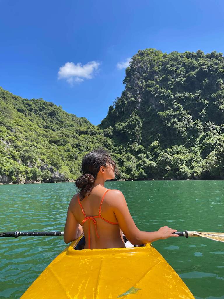 Solo female traveler kayaking during a day cruise around Ha Long Bay, La Hong Bay, and Cat Ba Vienam. She is wearing an orange bathing suit and sunglasses