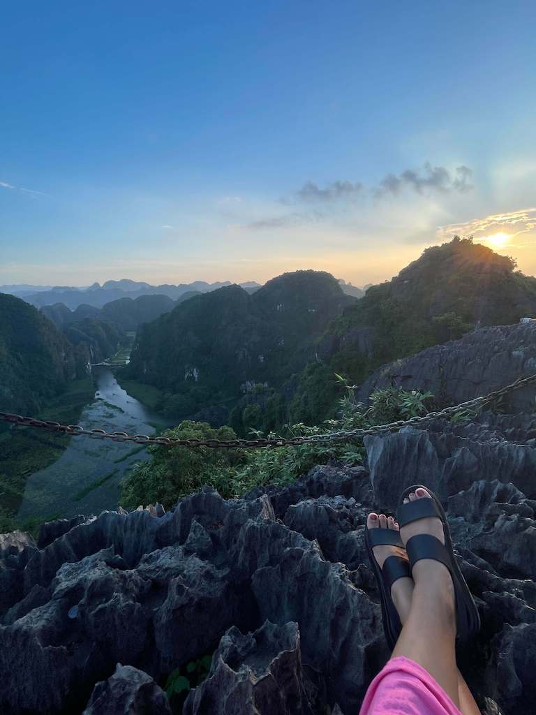 Backpacker sitting atop dragon mountain during sunset in Ninh Binh Vietnam, overlooking luscious greenery, cliffs and a river